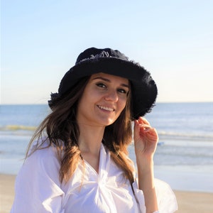May include: A woman wearing a white shirt and a black bucket hat with frayed edges. She is smiling and looking at the camera. The background is a beach with blue water and white sand.
