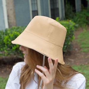 May include: A beige bucket hat with a white trim. The hat is being worn by a person with long, light brown hair.