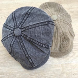 May include: Two newsboy caps. One is a faded denim blue with black stitching and a matching button on top. The other is a tan color with a matching button. Both caps are on a light wood surface.