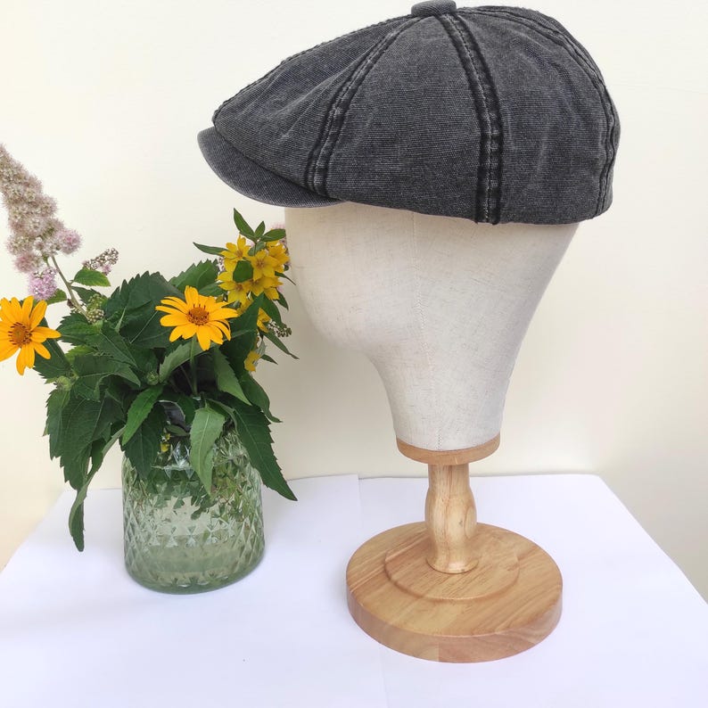 Peut inclure: Casquette de style gavroche gris fonc&eacute;, avec une couronne arrondie et une petite visi&egrave;re rigide. Le chapeau est pr&eacute;sent&eacute; sur une t&ecirc;te de mannequin avec un support en bois. Un vase de fleurs jaunes et de verdure est &agrave; gauche.