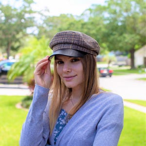 May include: A woman wearing a gray cardigan and a brown and black plaid newsboy cap with a black leather brim. She is smiling and looking at the camera.