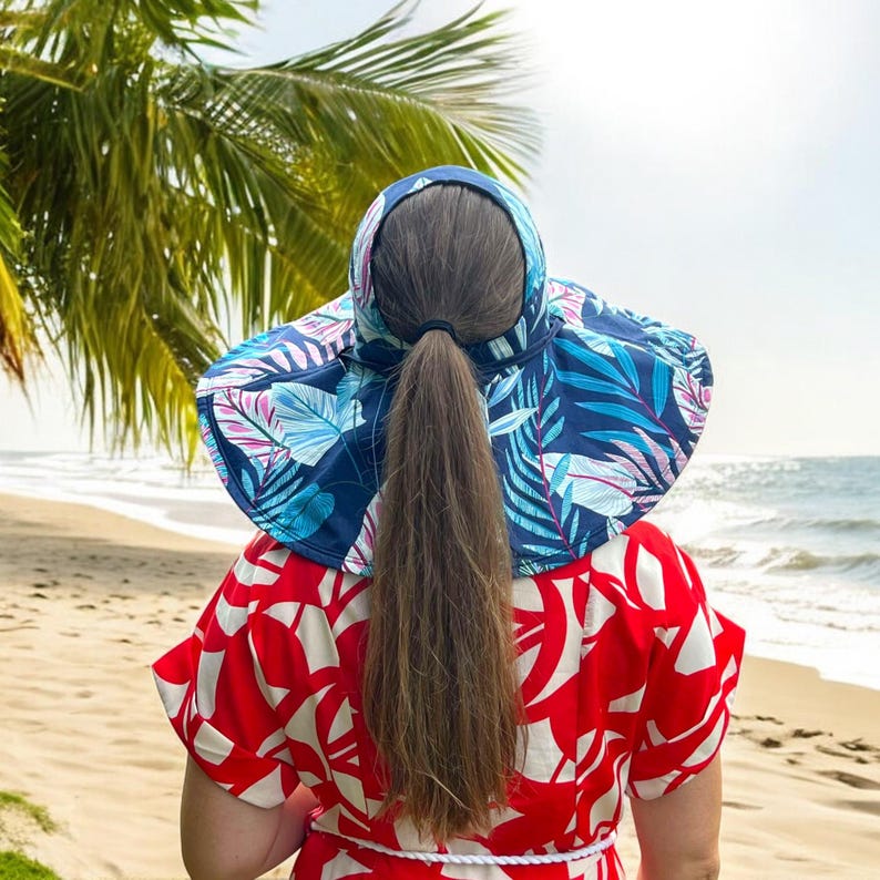 May include: A woman wearing a blue and pink floral sun hat with a wide brim and a neck flap. The hat is on a beach with a palm tree in the background.