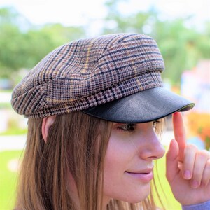 May include: A brown and black plaid newsboy cap with a black leather brim. The cap is worn by a person with long brown hair.
