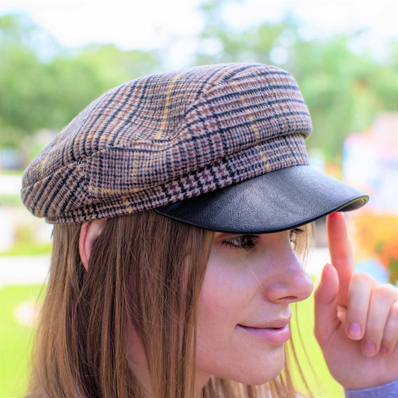 May include: A brown and black plaid newsboy cap with a black leather brim. The cap is worn by a person with long brown hair.