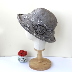 May include: A gray cloche hat with a floral pattern and a fabric rose accent. The hat is displayed on a mannequin head. The brim is approximately 3 inches wide. A small decorative pot with red roses is in the background.