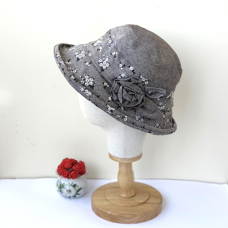 May include: A gray cloche hat with a floral pattern and a fabric rose accent. The hat is displayed on a mannequin head. The brim is approximately 3 inches wide. A small decorative pot with red roses is in the background.