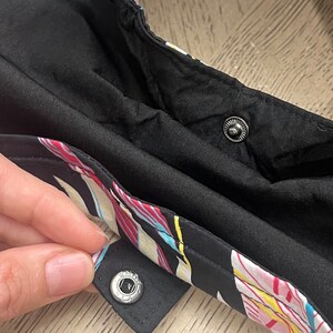 May include: Close-up of a black fabric with a colorful floral print. The fabric has two silver snap buttons.