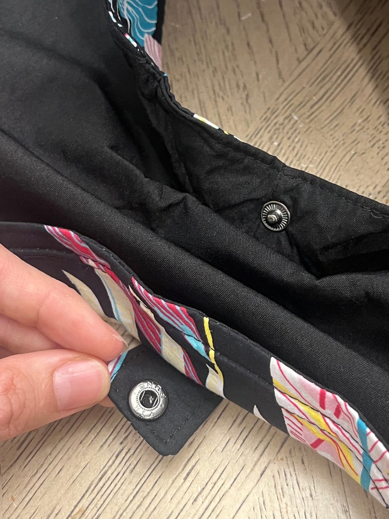May include: Close-up of a black fabric with a colorful floral print. The fabric has two silver snap buttons.