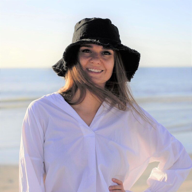 May include: A woman wearing a white button-down shirt with a black bucket hat. She is standing on a beach with the ocean in the background.