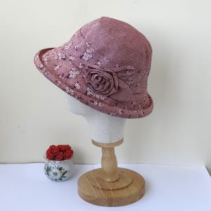 May include: A mauve cloche hat with a floral pattern and a fabric rose accent. The hat is displayed on a mannequin head and has a small brim. A small decorative pot with red roses is in the foreground.