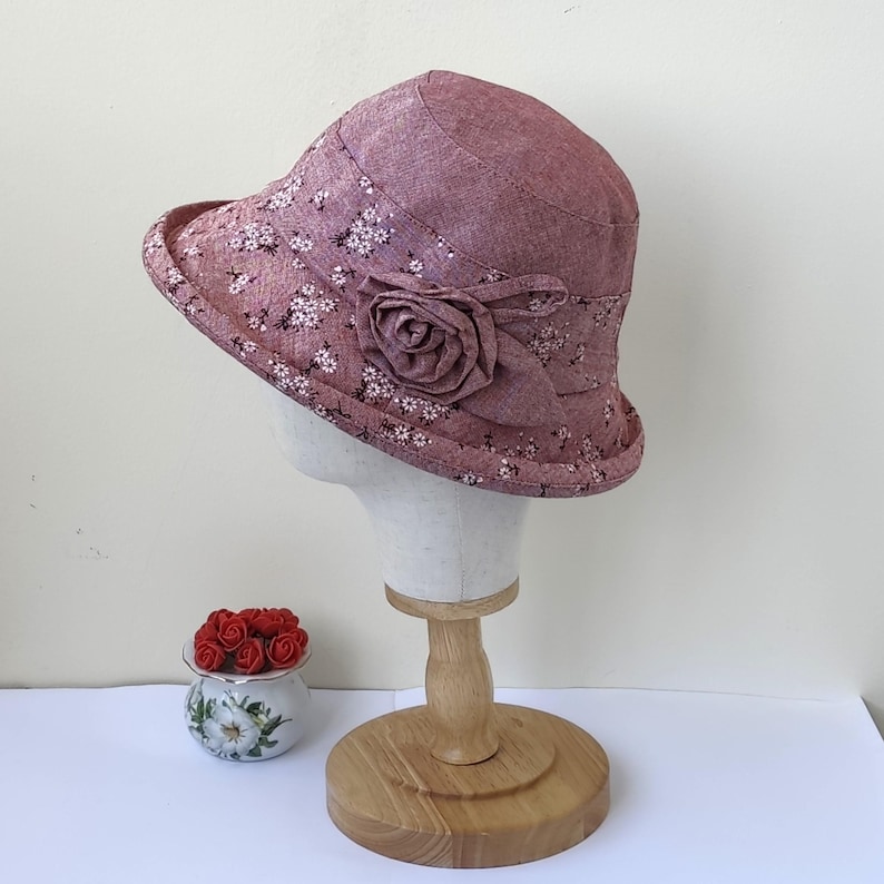 May include: A mauve cloche hat with a floral pattern and a fabric rose accent. The hat is displayed on a mannequin head and has a small brim. A small decorative pot with red roses is in the foreground.