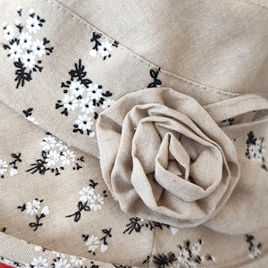 May include: A beige fabric hat with a floral pattern and a large, fabric rose accent. The hat features a repeating pattern of small white flowers with black stems and leaves. The rose is made of the same beige fabric.