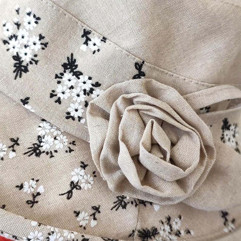 May include: A beige fabric hat with a floral pattern and a large, fabric rose accent. The hat features a repeating pattern of small white flowers with black stems and leaves. The rose is made of the same beige fabric.