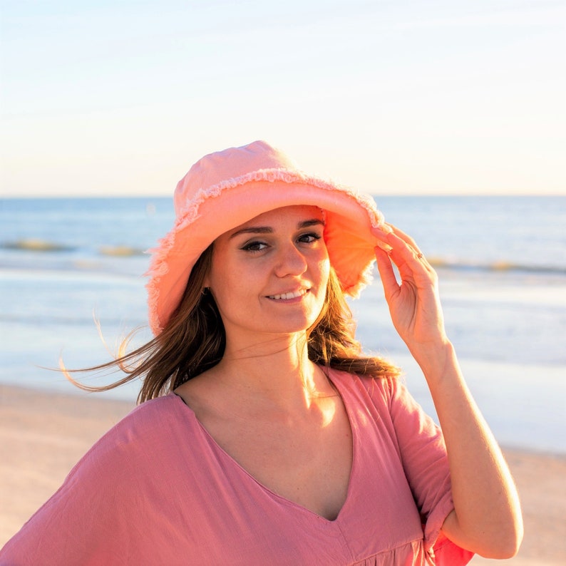 May include: A woman wearing a pink sun hat with a ruffled brim and a pink dress. She is standing on a beach with the ocean in the background.