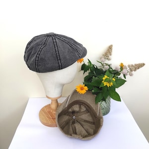 Peut inclure: Deux casquettes de style gavroche sont pr&eacute;sent&eacute;es sur une surface blanche. L'une est en tissu gris fonc&eacute; textur&eacute;, l'autre est de couleur beige. La casquette grise est sur une t&ecirc;te de mannequin. Un petit bouquet de fleurs jaunes et de verdure est en arri&egrave;re-plan.