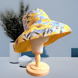 May include: A yellow sun hat with a floral print featuring blue, yellow, and white flowers. The hat is on a wooden display stand.