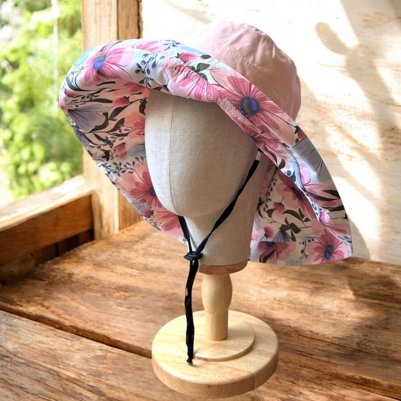 May include: A pink and white floral sun hat with a black adjustable chin strap. The hat is on a wooden display stand.