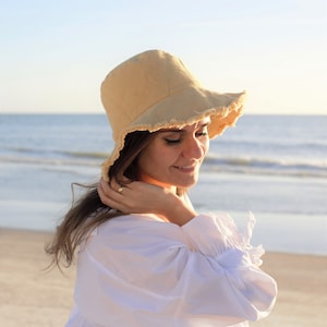 May include: A woman wearing a white shirt and a light beige bucket hat with frayed edges. The hat has a wide brim and is tilted down over her eyes. She is standing on a beach with the ocean in the background.