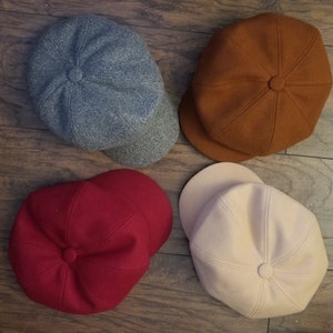 May include: Four newsboy caps in various colours: grey, brown, red, and cream. Each cap features a rounded crown and a small, stiff peak. The caps are made from a soft, textured fabric.