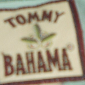 May include: A close-up of a Tommy Bahama label with a brown border and a white background. The label features a stylized green plant with a brown stem and a white flower.