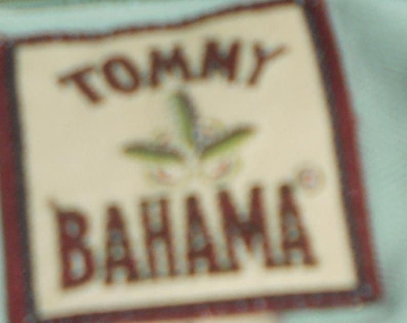 May include: A close-up of a Tommy Bahama label with a brown border and a white background. The label features a stylized green plant with a brown stem and a white flower.