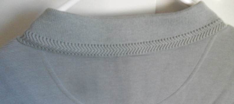 May include: A light gray polo shirt with a ribbed collar and a subtle textured pattern. The collar has a decorative stitching detail.