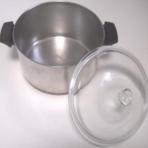 May include: A stainless steel cooking pot with black handles and a clear glass lid. The pot is round with a wide opening. The lid is off to the side, showing the interior of the pot. The pot is designed for cooking.