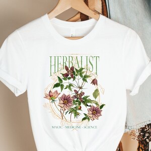 May include: White t-shirt with a floral design and the text "Herbalist: Magic - Medicine - Science".