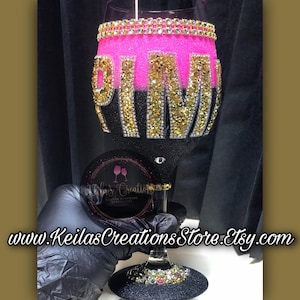 May include: A pink and black wine glass with the word "PINK" in gold glitter and rhinestones. The glass is decorated with rhinestones around the rim.