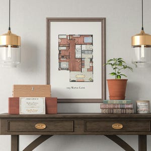 May include: A framed architectural floor plan print labeled "123 Maple Lane" sits on a dark wood console table. A small potted plant, books, and decorative boxes are also on the table. Two gold and glass pendant lights hang above.