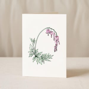 May include: A folded greeting card featuring a botanical illustration of a bleeding heart flower. The artwork showcases a curved stem with pink heart-shaped flowers and green leaves against a white background.