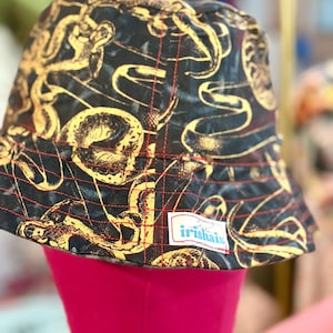 Może przedstawiać: Czarny kapelusz typu bucket z złotym wzorem węża i czerwonymi przeszyciami. Kapelusz ma małą białą etykietę z napisem "irishais". Kapelusz jest prezentowany na różowej głowie manekina.