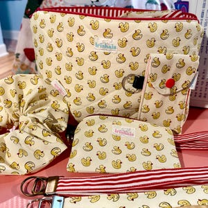 Könnte beinhalten: Eine gesteppte cremefarbene Tasche mit gelbem Entenmuster, rotem Reißverschluss und passendem Zubehör, darunter ein Haargummi, ein kleines Täschchen und zwei Schlüsselbänder mit rot-weißen Streifen. Jeder Artikel hat ein weißes Etikett mit dem Text irishais.