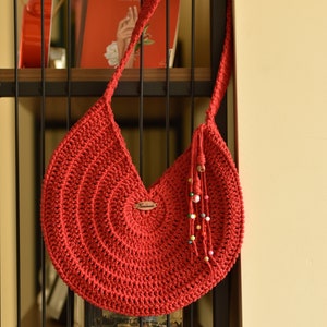 May include: A handmade red crochet shoulder bag with a circular design. The bag has a long strap and a decorative tassel with colorful beads. A small wooden tag with the word "Handmade" is attached to the bag.