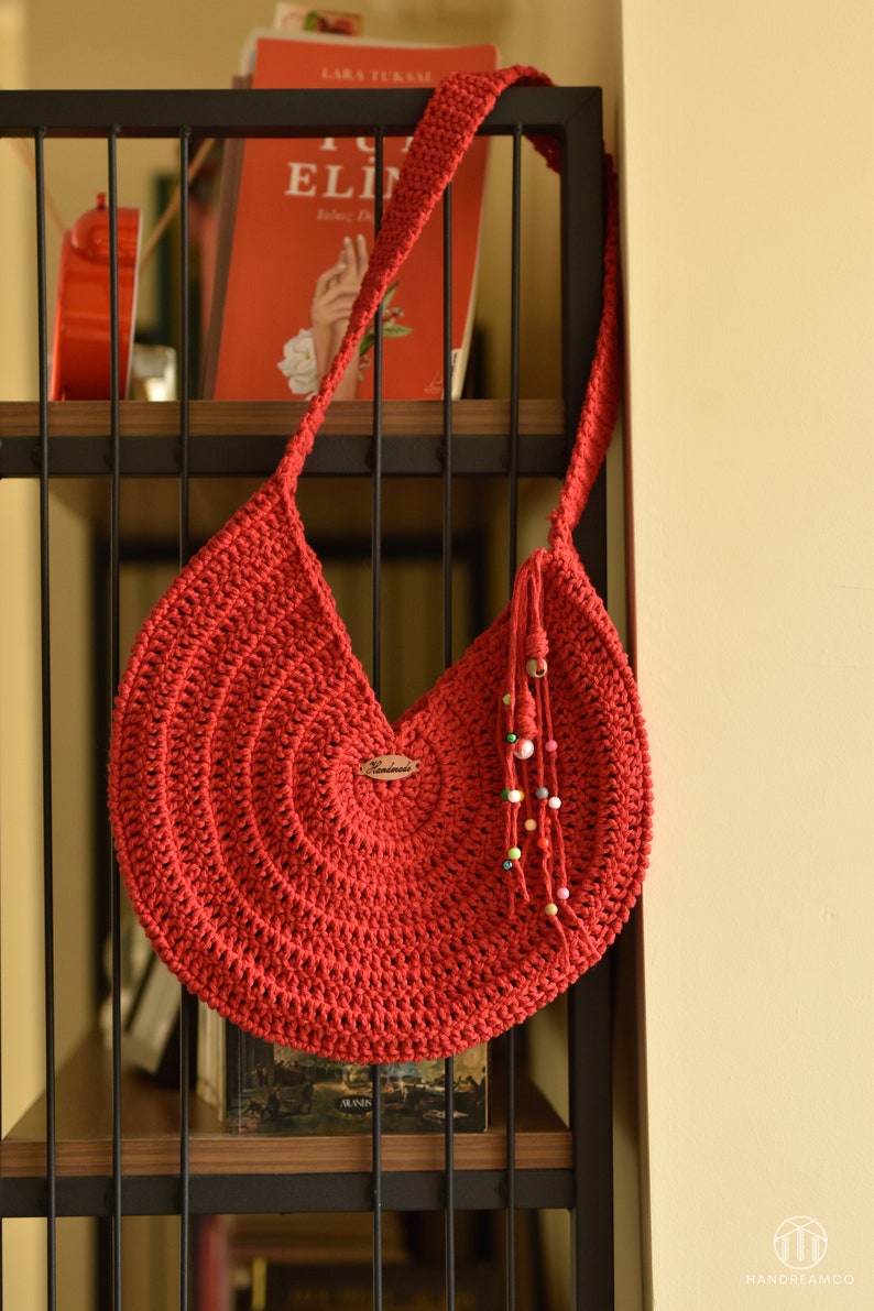 May include: A handmade red crochet shoulder bag with a circular design. The bag has a long strap and a decorative tassel with colorful beads. A small wooden tag with the word "Handmade" is attached to the bag.