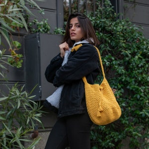 May include: A woman wearing a black denim jacket and black pants is standing in front of a wall with green vines. She is carrying a bright yellow crocheted tote bag with a drawstring closure.