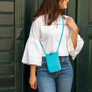 Puede incluir: Una mujer con una camiseta blanca y un bolso bandolera de crochet azul. El bolso tiene un peque&ntilde;o cierre de bot&oacute;n y una correa larga. La mujer est&aacute; de pie frente a una puerta de madera verde.