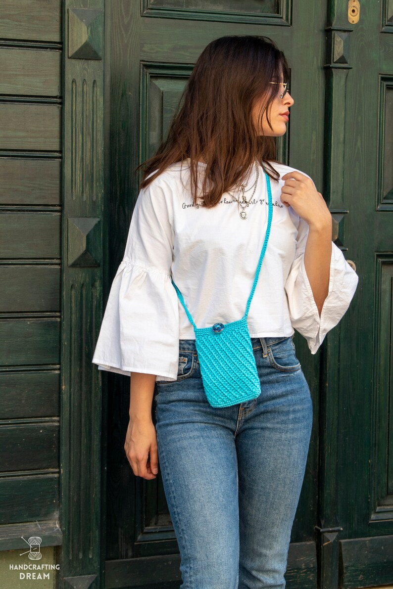 Puede incluir: Una mujer con una camiseta blanca y un bolso bandolera de crochet azul. El bolso tiene un peque&ntilde;o cierre de bot&oacute;n y una correa larga. La mujer est&aacute; de pie frente a una puerta de madera verde.