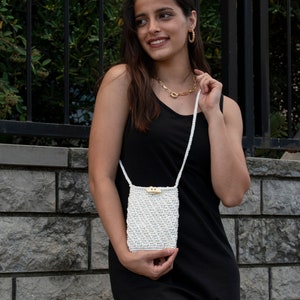Puede incluir: Un bolso bandolera de macram&eacute; blanco con cierre de bot&oacute;n de madera. El bolso lo lleva una persona que lleva un top negro de tirantes y un collar de oro.