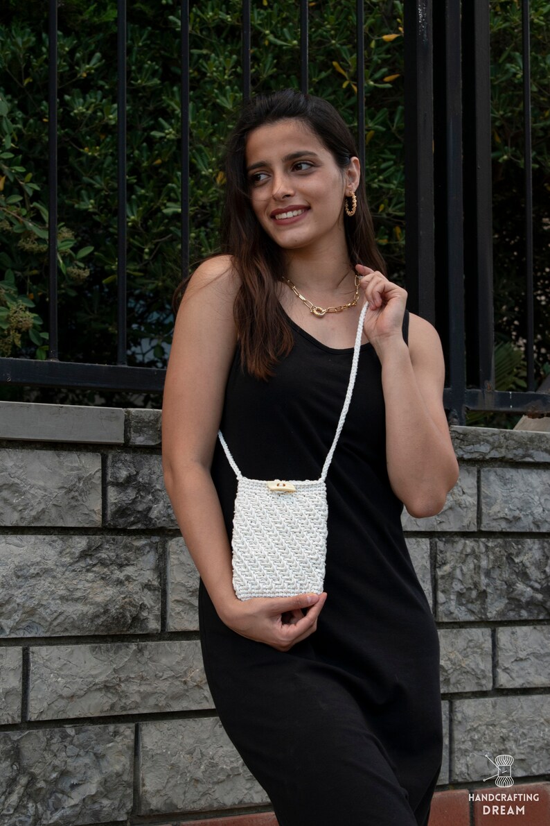 Puede incluir: Un bolso bandolera de macram&eacute; blanco con cierre de bot&oacute;n de madera. El bolso lo lleva una persona que lleva un top negro de tirantes y un collar de oro.