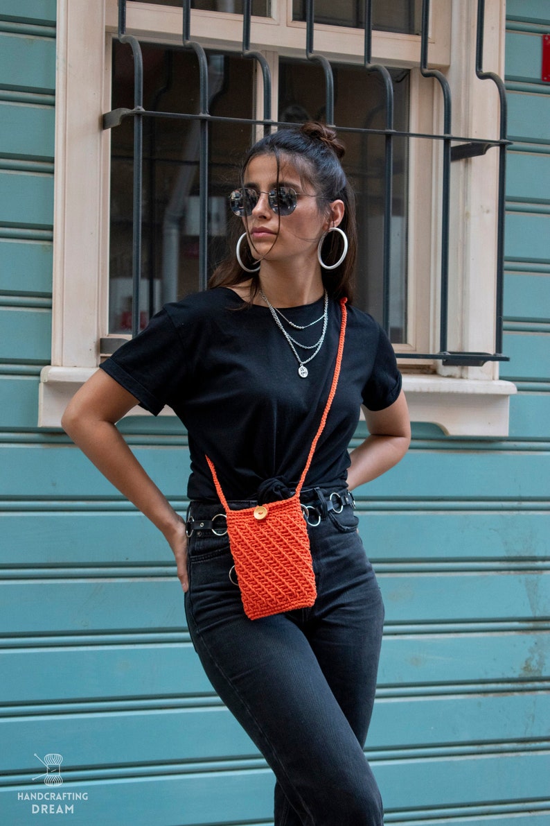 Puede incluir: Una mujer con una camiseta negra, pantalones vaqueros negros y un bolso bandolera de crochet naranja brillante con cierre de bot&oacute;n. Lleva gafas de sol y pendientes de aro plateados.