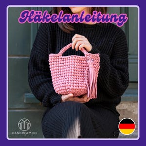 May include: A pink crocheted handbag with a tassel. The bag is being held by a person wearing a black sweater. The text "Häkelanleitung" is displayed at the top of the image.