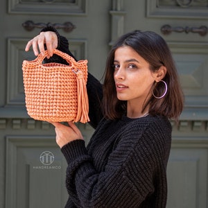 Pu&ograve; includere: Una borsa a mano in crochet color pesca con un pon pon. La borsa &egrave; tenuta da una persona che indossa un maglione nero.