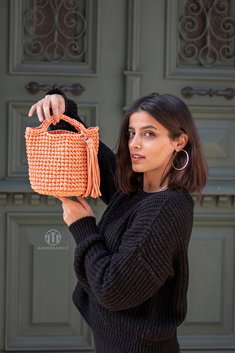 Pu&ograve; includere: Una borsa a mano in crochet color pesca con un pon pon. La borsa &egrave; tenuta da una persona che indossa un maglione nero.