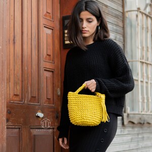 Pu&ograve; includere: Una donna con un maglione e una gonna neri tiene una piccola borsa di uncinetto gialla con un fiocco. La borsa &egrave; realizzata con filato spesso e ha un design semplice.