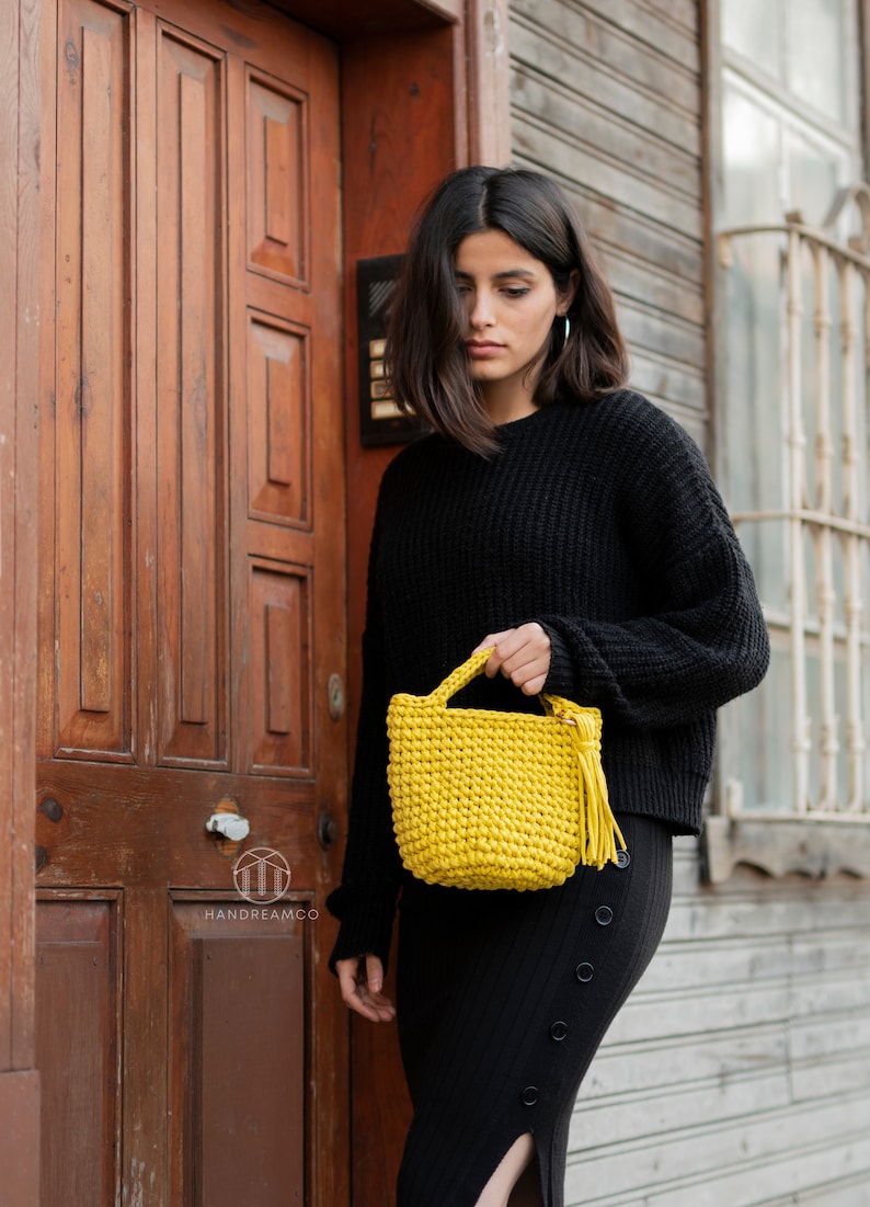 Pu&ograve; includere: Una donna con un maglione e una gonna neri tiene una piccola borsa di uncinetto gialla con un fiocco. La borsa &egrave; realizzata con filato spesso e ha un design semplice.