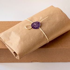 Puede incluir: Una caja de cart&oacute;n marr&oacute;n con un paquete de papel marr&oacute;n en la parte superior. El paquete est&aacute; atado con una cuerda y tiene una pegatina morada que dice "Handcrafting Dream".