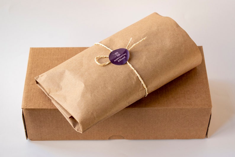 Puede incluir: Una caja de cart&oacute;n marr&oacute;n con un paquete de papel marr&oacute;n en la parte superior. El paquete est&aacute; atado con una cuerda y tiene una pegatina morada que dice "Handcrafting Dream".