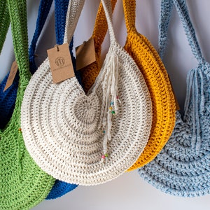 May include: A collection of handmade crochet bags in various colors, including green, blue, yellow, and off-white. Each bag has a round shape with a long strap and a tag. The off-white bag has a tassel with colorful beads.