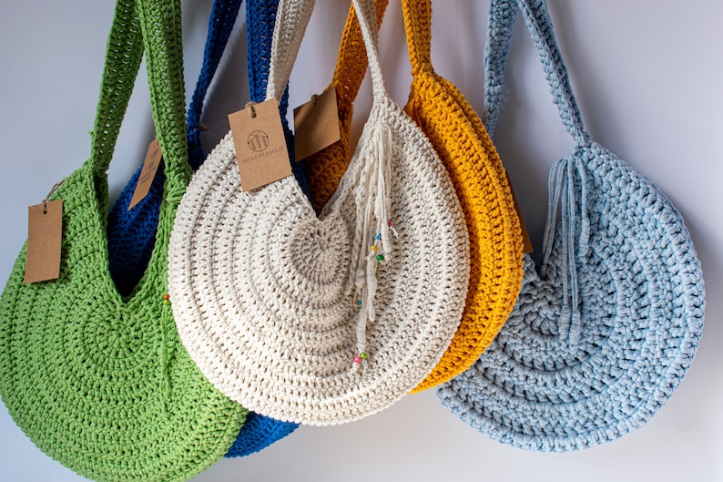 May include: A collection of handmade crochet bags in various colors, including green, blue, yellow, and off-white. Each bag has a round shape with a long strap and a tag. The off-white bag has a tassel with colorful beads.